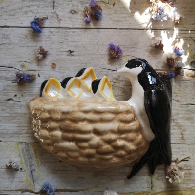 Figura de cerâmica de um ninho com ovos amarelos e um pássaro preto e branco sobre fundo de madeira e flores secas