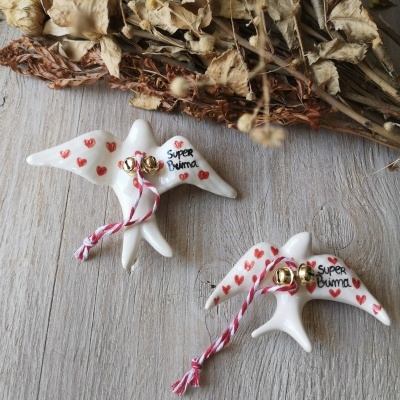 Figuras de pássaros em cerâmica branca com corações vermelhos, campainhas douradas e texto 'Super Prima' num fundo de madeira clara e folhas secas