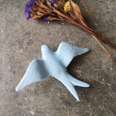 Pequena figura decorativa azul em forma de andorinha sobre superfície cinzenta com ramo seco e flores coloridas