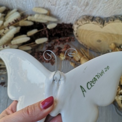 Borboleta decorativa em cerâmica branca com antenas de arame e texto A CREATIVA 26, mão segurando, fundo com flores secas e madeira
