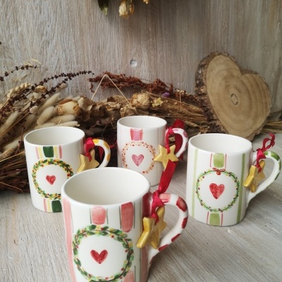 Conjunto de quatro canecas decoradas com riscas e coroas com corações vermelhos, cada uma com uma fita vermelha e estrela dourada, sobre mesa de madeira com flores secas e placa de madeira em forma de coração.