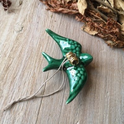 Pássaro decorativo verde em cerâmica com sinos dourados e cordão sobre superfície de madeira
