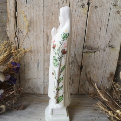 Estátua religiosa branca com flores pintadas, base quadrada, madeira rústica e plantas secas ao redor.