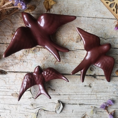 Três andorinhas decorativas em cerâmica marrom sobre madeira clara com flores secas