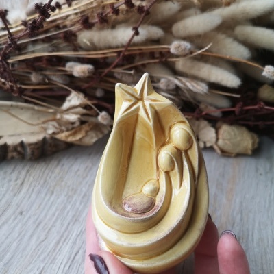 Escultura cerâmica amarela da Sagrada Família com estrela, mão segurando e ramos secos ao fundo