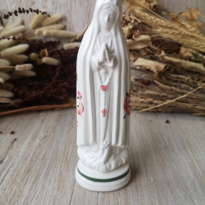 Estatueta branca de Nossa Senhora com terço e flores pintadas, sobre madeira clara com plantas secas ao fundo