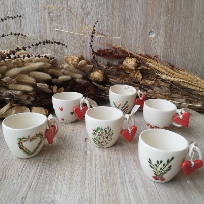 Conjunto de chávenas de cerâmica branca com decorações florais e corações vermelhos em cordão, sobre madeira clara e flores secas ao fundo.