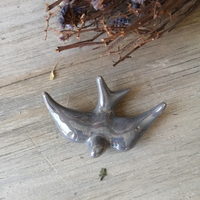 Peça decorativa prateada em forma de pássaro sobre madeira clara com ramo seco e flores