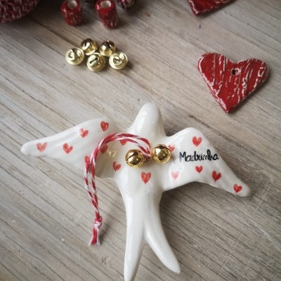 Pássaro decorativo branco com corações vermelhos, sinos dourados, texto 'Madrinha' e botões e corações vermelhos ao fundo sobre madeira clara