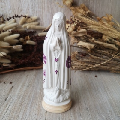 Estátua branca de Nossa Senhora com detalhes roxos em rosário e flores, sobre base creme e fundo de decoração seca