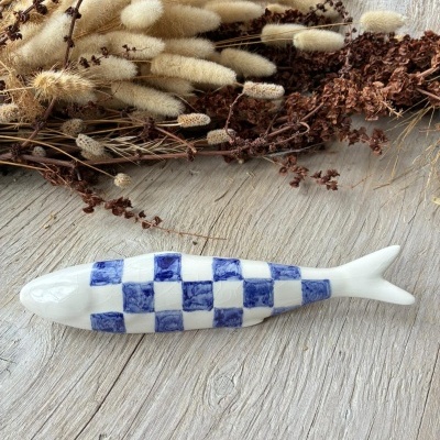 Peixe decorativo cerâmico branco com padrão xadrez azul ao lado de ramos secos sobre madeira.