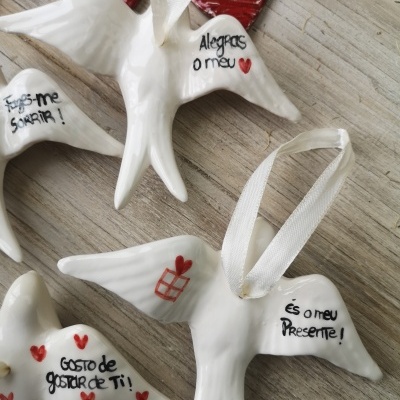 Ornamentos em forma de pássaros brancos com fitas e inscrições em português, sobre fundo de madeira clara com corações vermelhos.