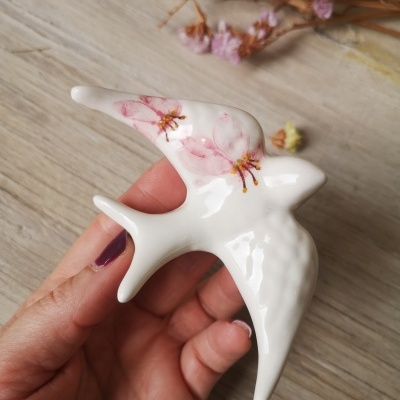 Puxador decorativo branco em forma de pássaro com flores rosas, mãos segurando, fundo madeira clara