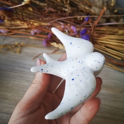 Peça decorativa branca em forma de pássaro com pintas azuis sendo segurada à frente de flores secas