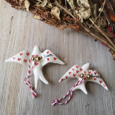 Ornamentos de cerâmica em forma de pássaros brancos com corações vermelhos e a palavra Primos, fitas vermelhas e sinos dourados sobre madeira clara.