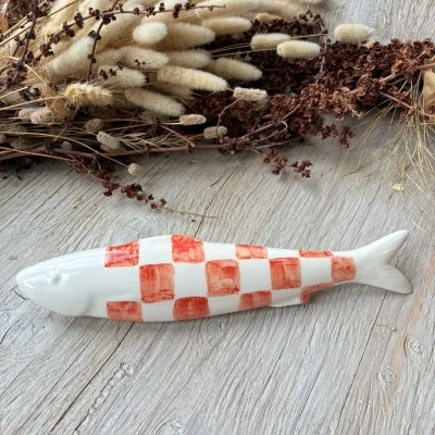 Peixe decorativo de cerâmica branca com quadrados vermelhos em fundo de madeira clara e ramos secos