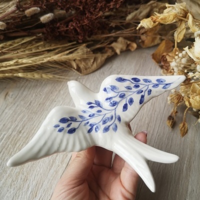 Figura cerâmica branca de andorinha com folhas azuis, mão a segurar e flores secas no fundo