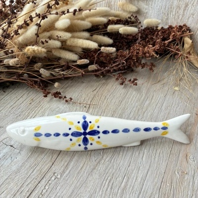 Figura decorativa de peixe em cerâmica branca com pintura azul e amarela junto a flores secas sobre superfície de madeira clara.