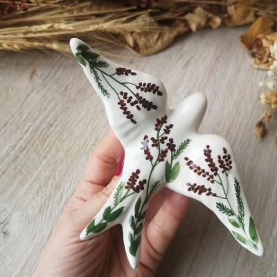 Pássaro decorativo cerâmico branco com pintura floral verde e castanha em fundo de madeira e flores secas