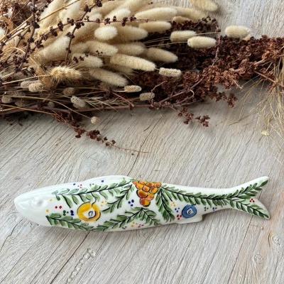 Figura cerâmica em forma de peixe com pintura floral colorida sobre mesa de madeira clara e flores secas ao fundo.
