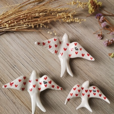Três figuras de pássaros em cerâmica branca com corações vermelhos e a palavra 'Tio', sobre madeira clara com ramo seco