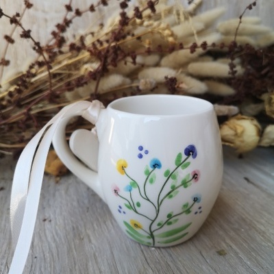Caneca branca com pintura colorida de flores e folhas, com fita branca na pega, em cima de madeira cinza e flores secas ao fundo