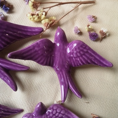 Figuras decorativas de pássaros roxos em cerâmica em tecido bege com flores secas.