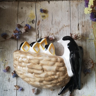 Objeto decorativo em cerâmica com ninho e passarinho preto e branco sobre fundo de madeira e flores secas