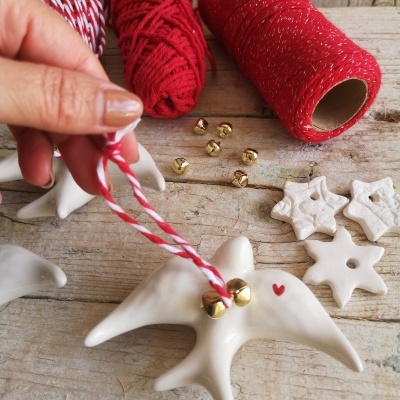 Decoração de passarinho branco em cerâmica com sinos dourados e fio vermelho e branco sobre mesa de madeira com materiais de artesanato.