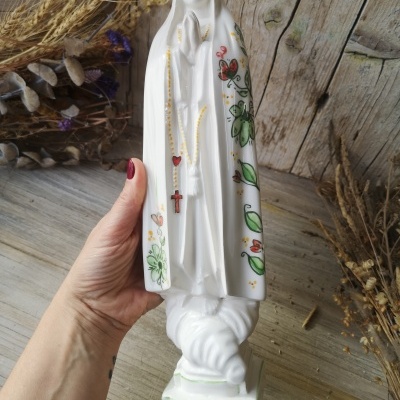 Estátua branca de porcelana de figura religiosa com desenhos florais e terço colorido.