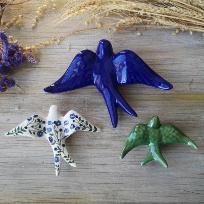 Broches em forma de andorinhas em azul, branco com flores e verde, sobre superfície de madeira clara com plantas secas