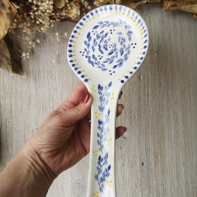 Colher de cerâmica branca decorada com padrão azul e amarelo, segurada à mão sobre fundo de madeira e flores secas.