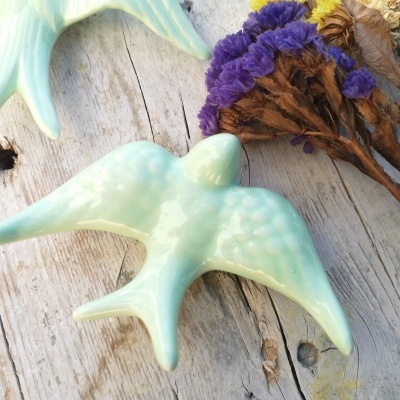 Pássaro decorativo azul claro em cerâmica com flores secas ao lado, sobre madeira clara