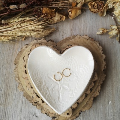 Brincos de argola dourados em prato de cerâmica branco em forma de coração com flores secas ao fundo