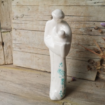 Estátua decorativa branca de cerâmica com detalhes verdes e azuis, sobre madeira clara com flores secas ao lado