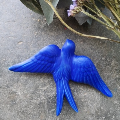 Figura decorativa azul em forma de pássaro sobre superfície cinzenta com folhas e flores secas ao fundo