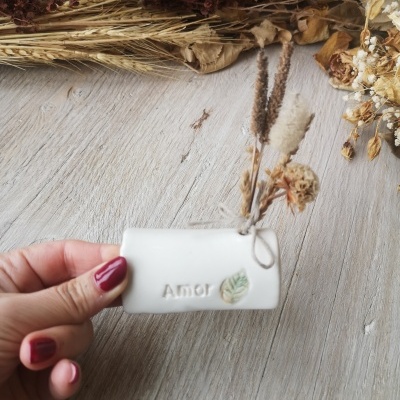 Vaso branco cerâmico pequeno com palavra Amor e folhas, cheio de ramos secos, mão segurando sobre mesa de madeira.