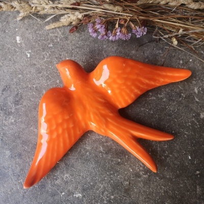 Pássaro decorativo em cerâmica laranja sobre superfície cinzenta com elementos naturais secos ao fundo