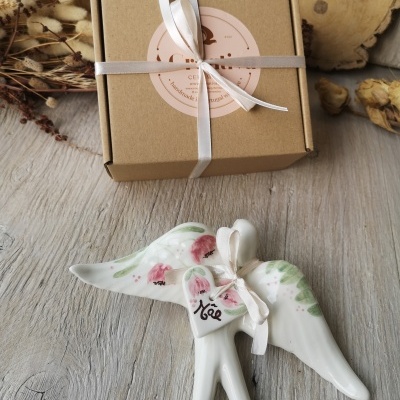 Estatueta branca de anjo em cerâmica pintada com fita e etiqueta ao lado de caixa de presente