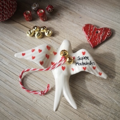 Pássaro decorativo branco com corações vermelhos e texto 'SUPER Madrinha' com sinos dourados em superfície de madeira