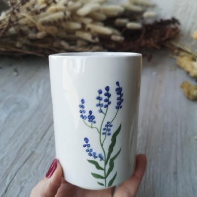 Caneca branca com flores azuis e folhas verdes segurada à mão, ramos secos ao fundo
