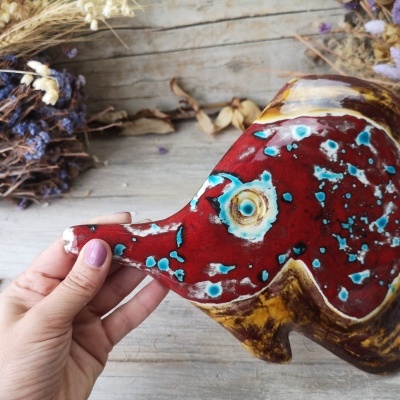 Escultura de peixe esmaltada em vermelho, azul e amarelo segurada por mão feminina sobre madeira cinzenta com ramos secos