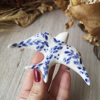 Figura de cerâmica em forma de pássaro com folhas azuis pintadas, segurada por mão feminina