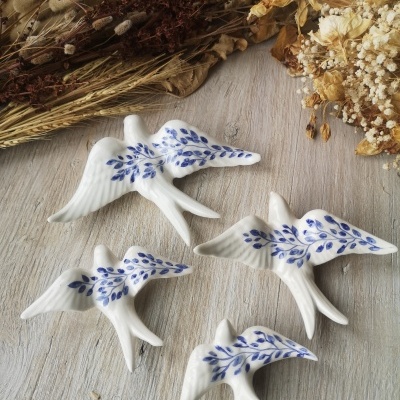 Figuras decorativas cerâmica branca aves com folhas azuis sobre mesa madeira clara com flores secas