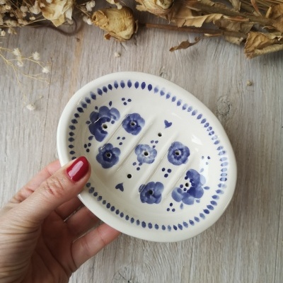 Prato de sabonete cerâmico branco com pintura azul floral e pequenos furos, mão segurando, sobre madeira clara, flores secas ao fundo.
