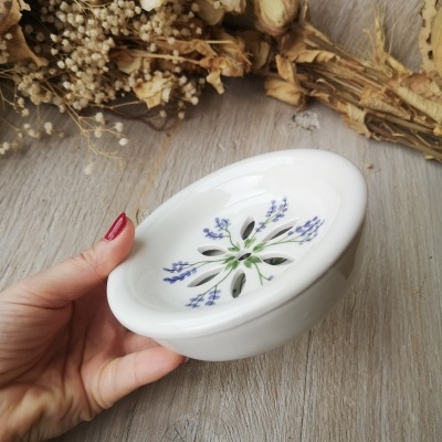 Saboneteira branca de cerâmica com padrão floral lilás e verde, mão segurando, ramos secos ao fundo.