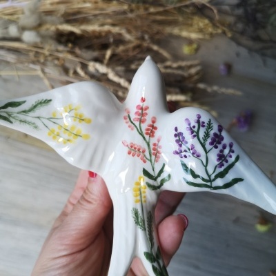 Figura cerâmica branca em forma de pássaro com flores pintadas, segurada por mão com unhas vermelhas