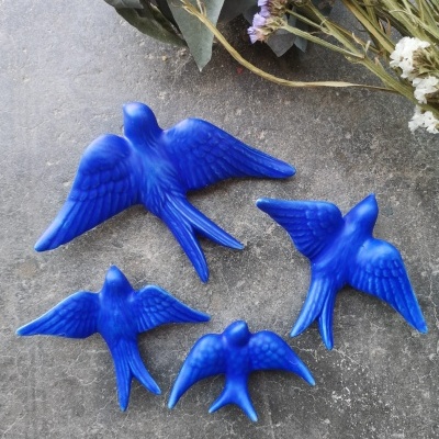 Quatro pássaros decorativos azuis de vários tamanhos sobre superfície cinza com flores secas ao fundo