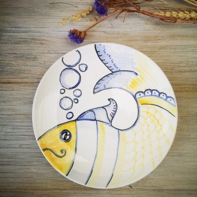Prato de cerâmica com desenho de peixe amarelo e azul sobre mesa de madeira com flores secas
