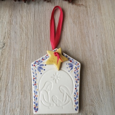 Enfeite de Natal em cerâmica branca com relevo da Natividade e fita vermelha.